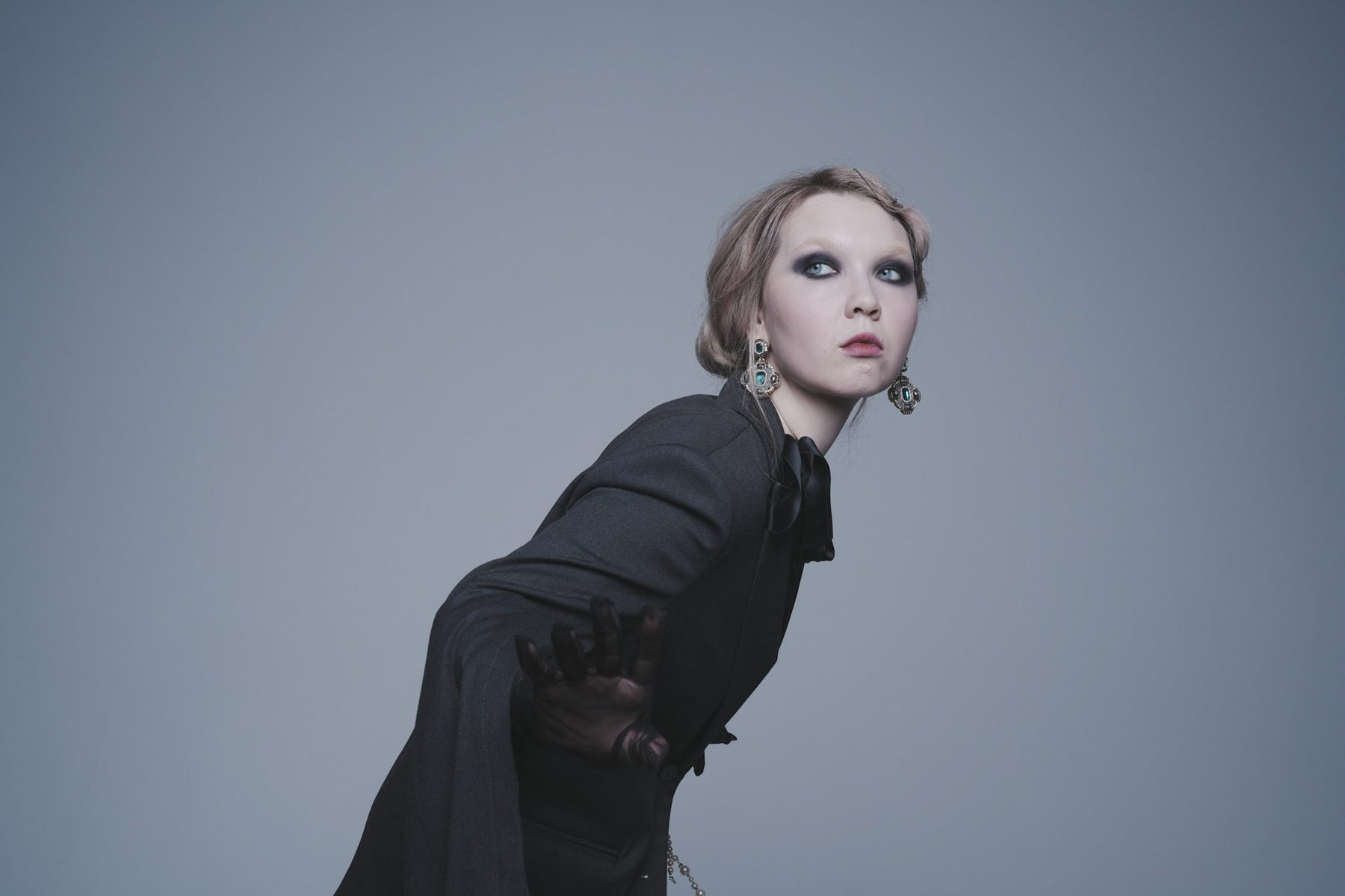 Editorial portrait in black tailoring on a steel-blue backdrop